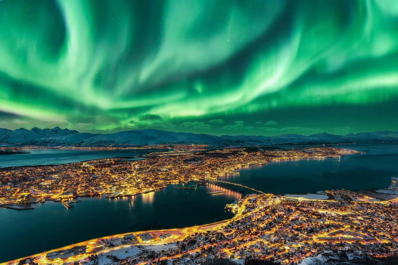 Aurora Borealis dancing over Tromso Urban Skyline. Tromso Panorama and winter night landscape with Northern Lights (Aurora Polaris) on a cold night, with starry sky, polar lights and snowcapped mountains on Kvaloya Island in the background. Cityscape with Brightly Street Light of the Arctic Capital, Northern Norway