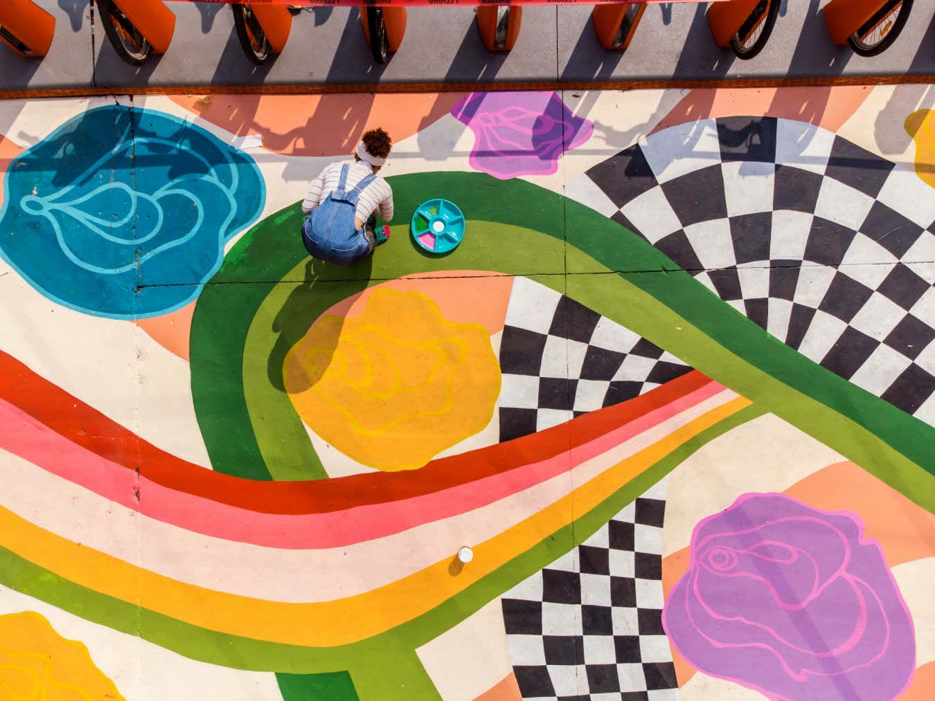 Young Latin woman artist painting sidewalk mural . She is dressed in casual work clothes. Exterior of public sidewalk in downtown of large North American City. Drone point of view.