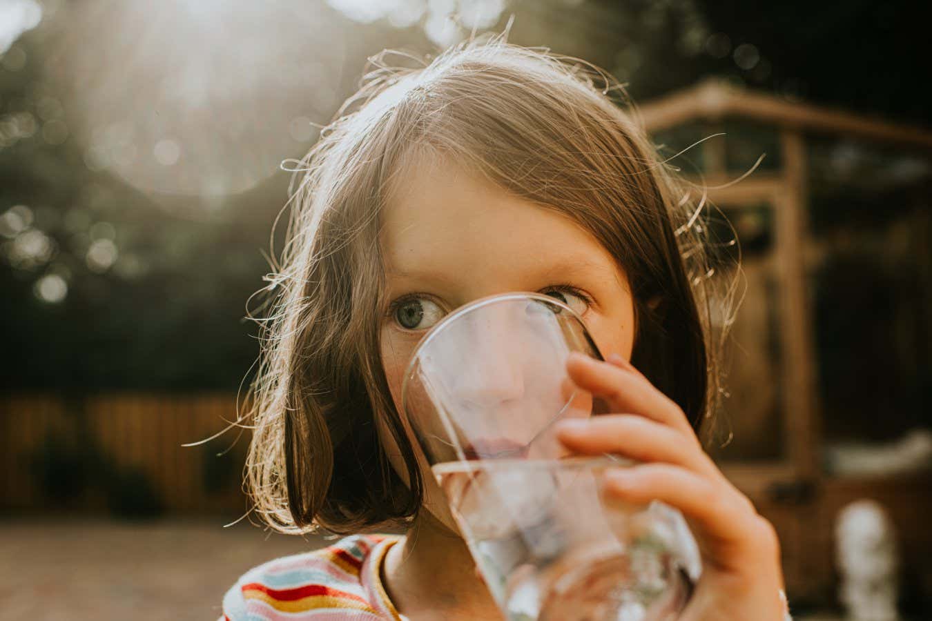 A cute young girl holds a transparent glass containing water with ice. She stands in a sunny garden and enjoys drinking the refreshing liquid. Space for copy.